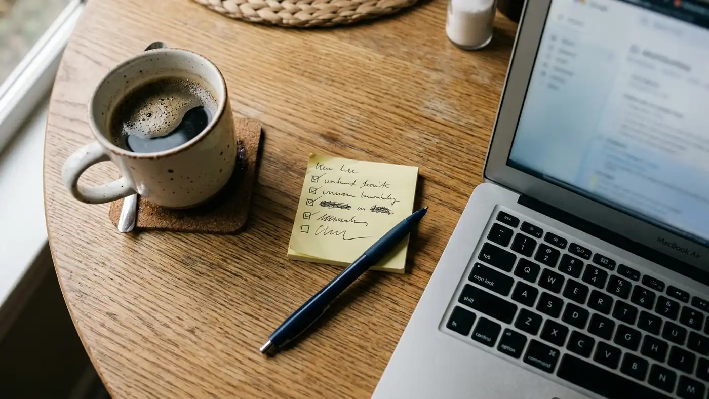 A handwritten decision checklist on a sticky note beside a laptop and coffee on a kitchen table